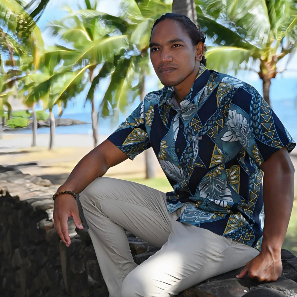 Homme portant une chemise tahitienne bleu à motifs polynésiens, assis devant des palmiers.