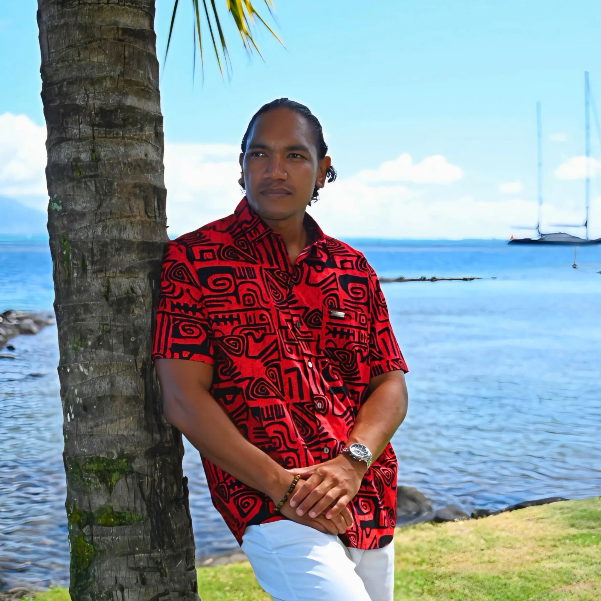 Homme portant une chemise tahitienne rouge et noirs à tatouages polynésiens, près d’un palmier en bord de mer